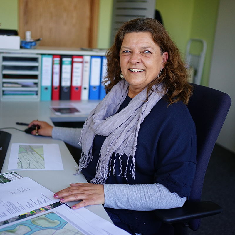 Frau mit dunkelblauem Pullover und hellgrauem Schal sitzt an Schreibtisch mit Papieren und Ordnern im Hintergrund.