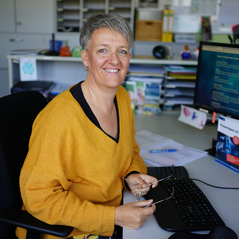 Person mit kurzem grauem Haar und gelbem Pullover sitzt an einem Schreibtisch mit Tastatur, Monitor und Unterlagen und hält eine Brille in der Hand.