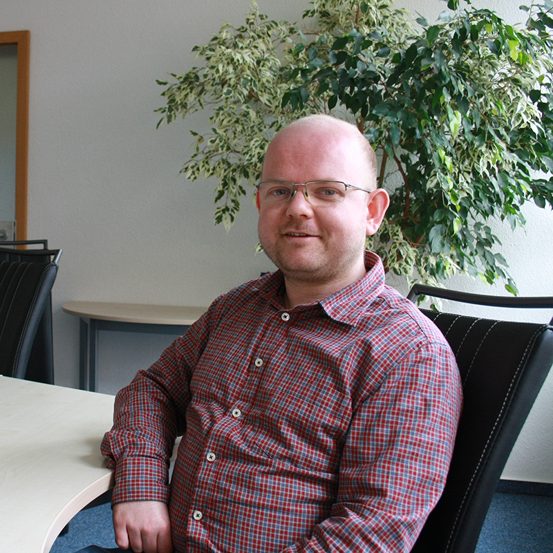 Mann mit kurz geschorenem Haar trägt ein rot-kariertes Hemd und sitzt an einem Tisch in einem Büro mit Pflanzen im Hintergrund.