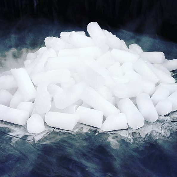 Haufen weißer, zylindrischer Trockeneis-Nuggets auf dunklem Untergrund mit Nebelbildung.