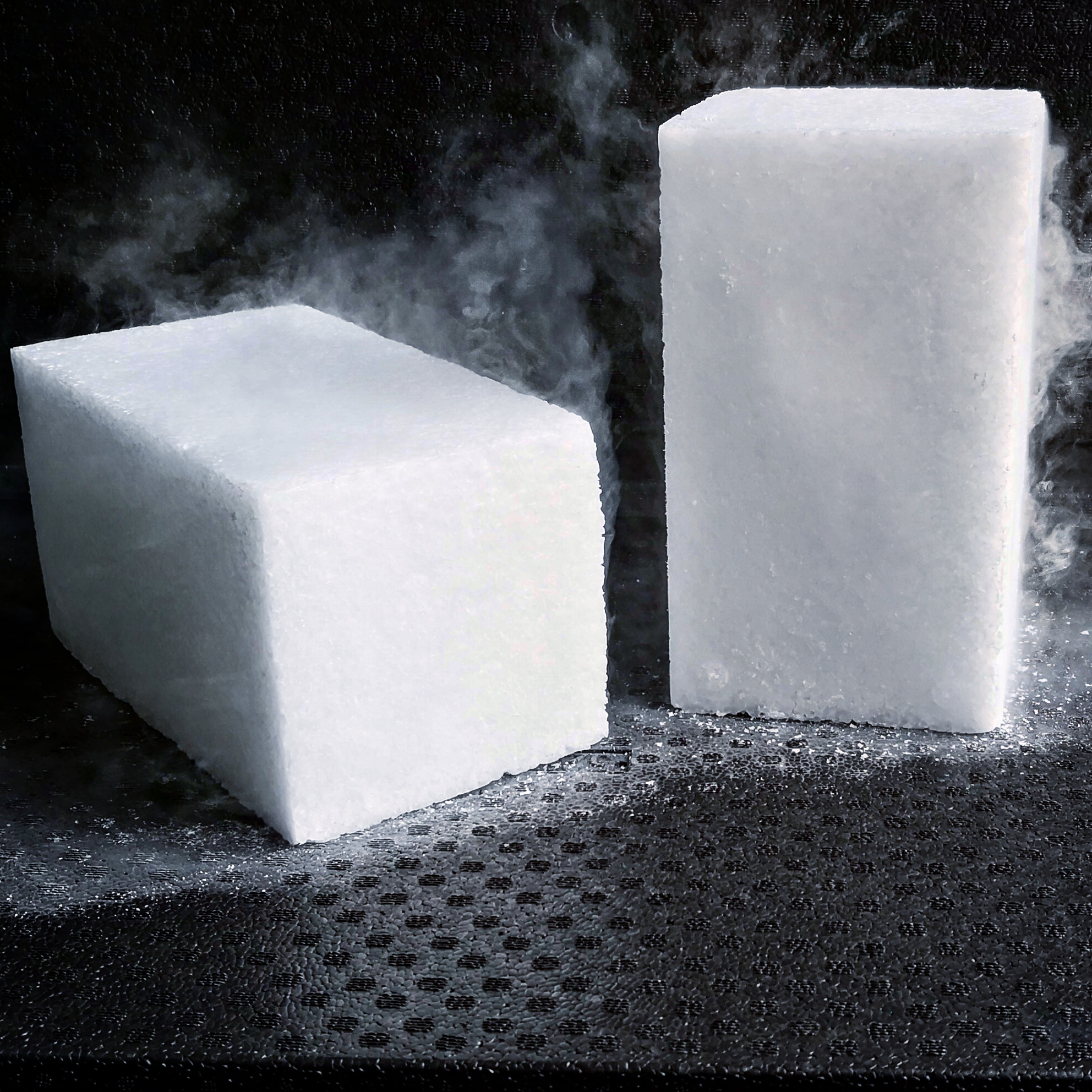 Zwei weiße Trockeneisblöcke in rechteckiger Form mit aufsteigendem Nebel auf dunklem Untergrund.