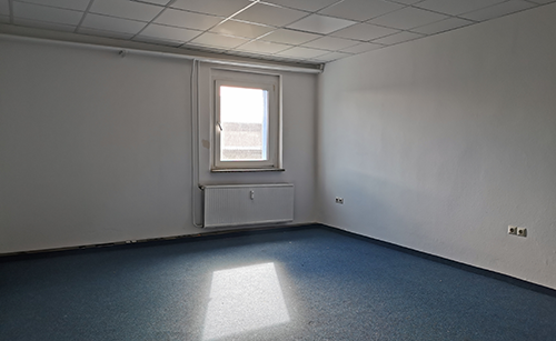 Leerer Büroraum mit blauem Teppichboden, weiß gestrichenen Wänden, einem Fenster mit Blick auf eine Betonwand und einer Heizung darunter.