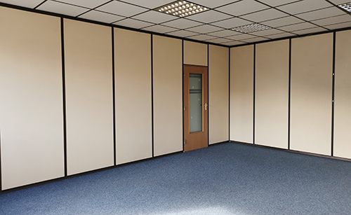 Leerer Büroraum mit blauer Teppichbodenfläche, hellen Trennwänden und einer Holztür mit Glaseinsatz.