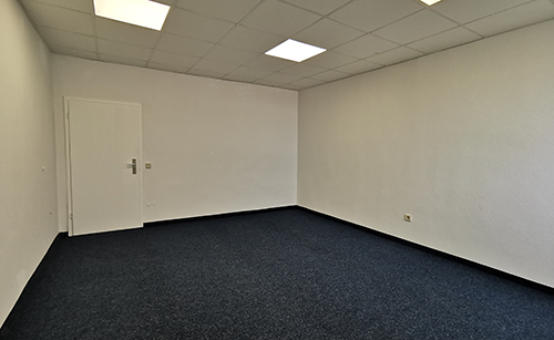 Leerer Büroraum mit weiß gestrichenen Wänden, dunkelblauem Teppichboden, weißer Tür und Deckenbeleuchtung.