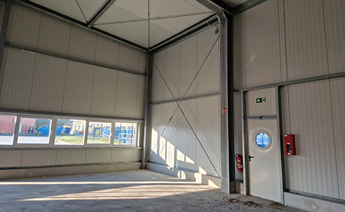 Innenansicht einer leeren Lagerhalle mit hohen Wänden, Fenstern, einer weißen Tür mit rundem Fenster und einem Feuerlöscher an der Wand.
