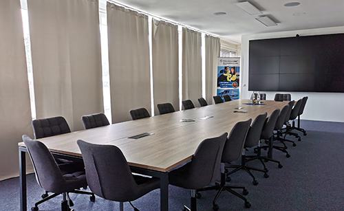 Moderner Tagungsraum mit langem rechteckigem Holztisch, mehreren schwarzen Bürostühlen und großen Fenstern mit geschlossenen Vorhängen.