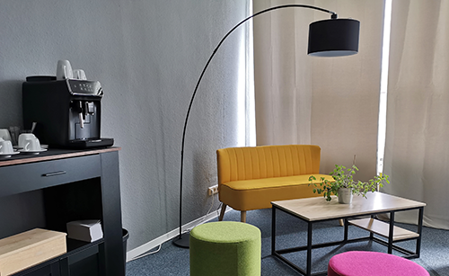 Gelbe zweisitzige Couch, bunter Hocker, schwarzer Bogenlampenschirm, Kaffeepadmaschine auf schwarzem Sideboard in Loungeecke.