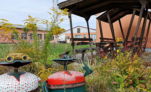 Zwei bunte Froschskulpturen mit großen Augen stehen in einem Garten mit gelben Blumen vor einem überdachten Sitzbereich und Gebäuden.