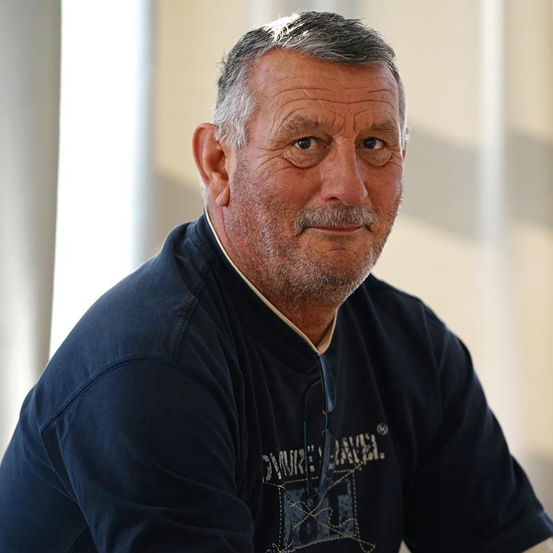 Mann mit grauen Haaren und Bart trägt ein dunkles T-Shirt mit hellem Aufdruck und sitzt vor einem hellen Hintergrund.