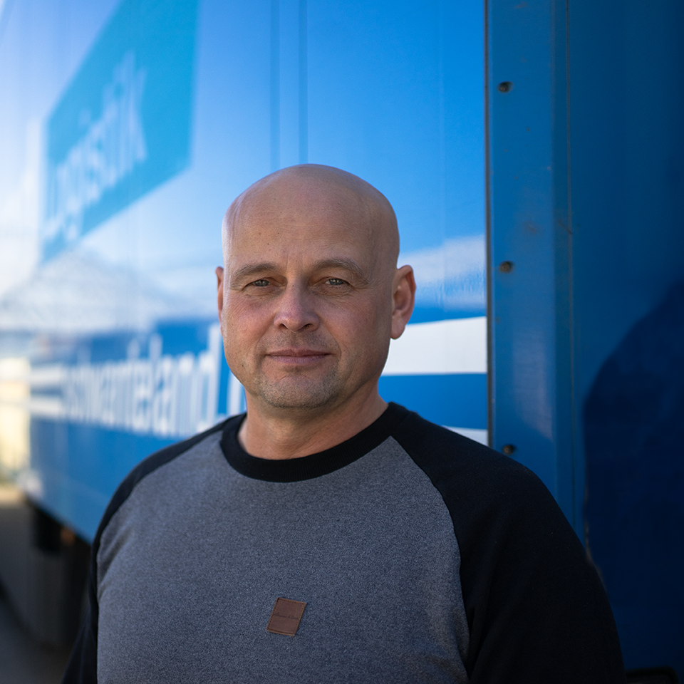 Mann mit grauem Pullover mit schwarzen Ärmeln steht vor einem blauen Lkw mit weißer Schrift.