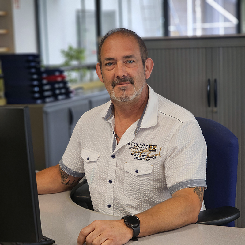 Mann mit kurzärmligem, weißem Hemd mit Brusttaschen und Aufdruck sitzt an Schreibtisch mit Computer in Büro.