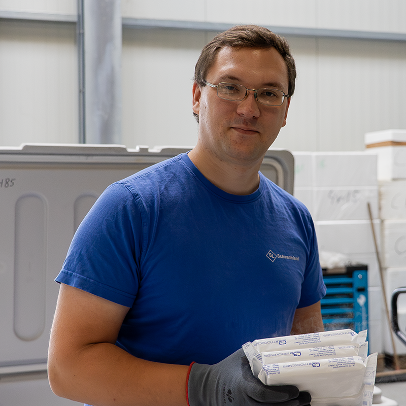 Mann in blauem T-Shirt mit Logo hält mehrere weiße Trockeneis-Pakete in grauen Handschuhen in einer Lagerhalle.