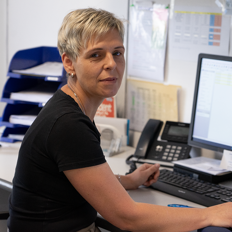 Person mit kurzem blonden Haar sitzt an einem Schreibtisch und arbeitet an einem Computer mit Tastatur und Telefon daneben.
