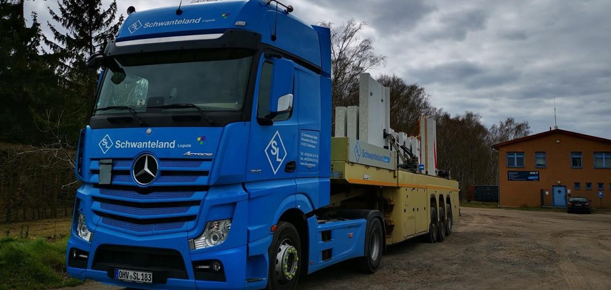Blauer Mercedes-Lkw mit Aufschrift 'Schwanteland' transportiert mehrere große Betonfertigteile auf gelbem Anhänger auf unbefestigtem Weg.