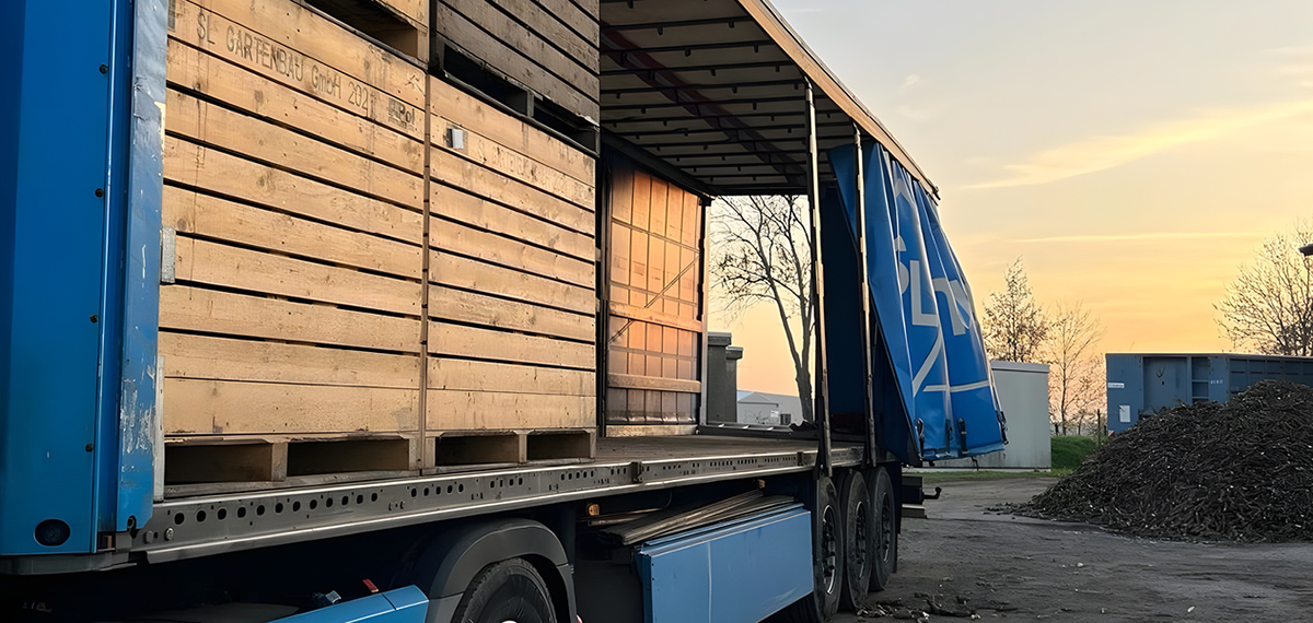 Blauer Lkw-Anhänger mit offener Plane, beladen mit gestapelten Holzkisten, im Sonnenuntergang auf einem Industriehof.