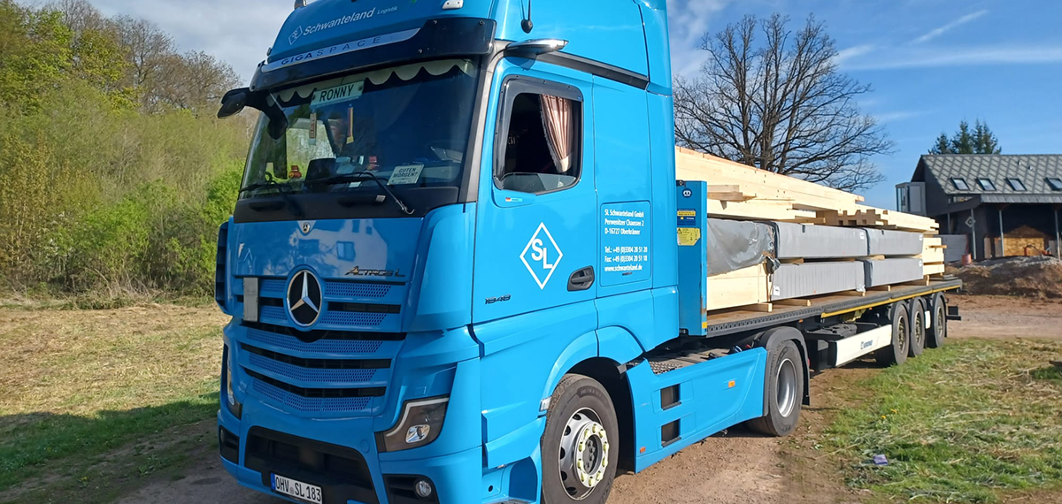 Blauer Mercedes-Lkw mit offenem Fenster, beladen mit langen Holzbalken, steht auf einem Feldweg vor Bäumen und einem Haus.