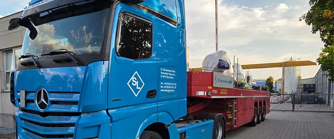 Blauer Mercedes-Lkw mit SL-Logo auf der Tür, beladen mit mehreren großen, weiß abgedeckten Skulpturen auf rotem Anhänger.