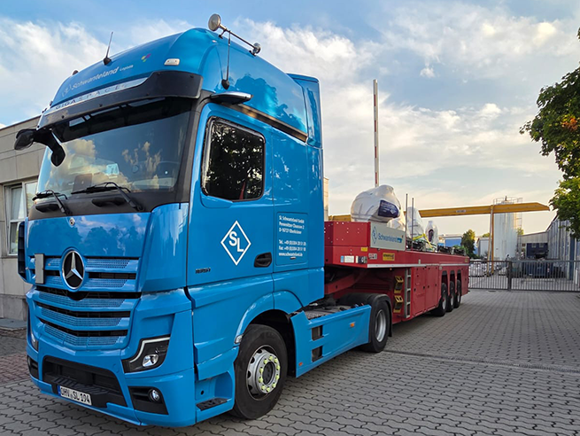 Blauer Mercedes-Lkw mit SL-Logo auf der Tür, beladen mit mehreren großen, weiß abgedeckten Skulpturen auf rotem Anhänger.