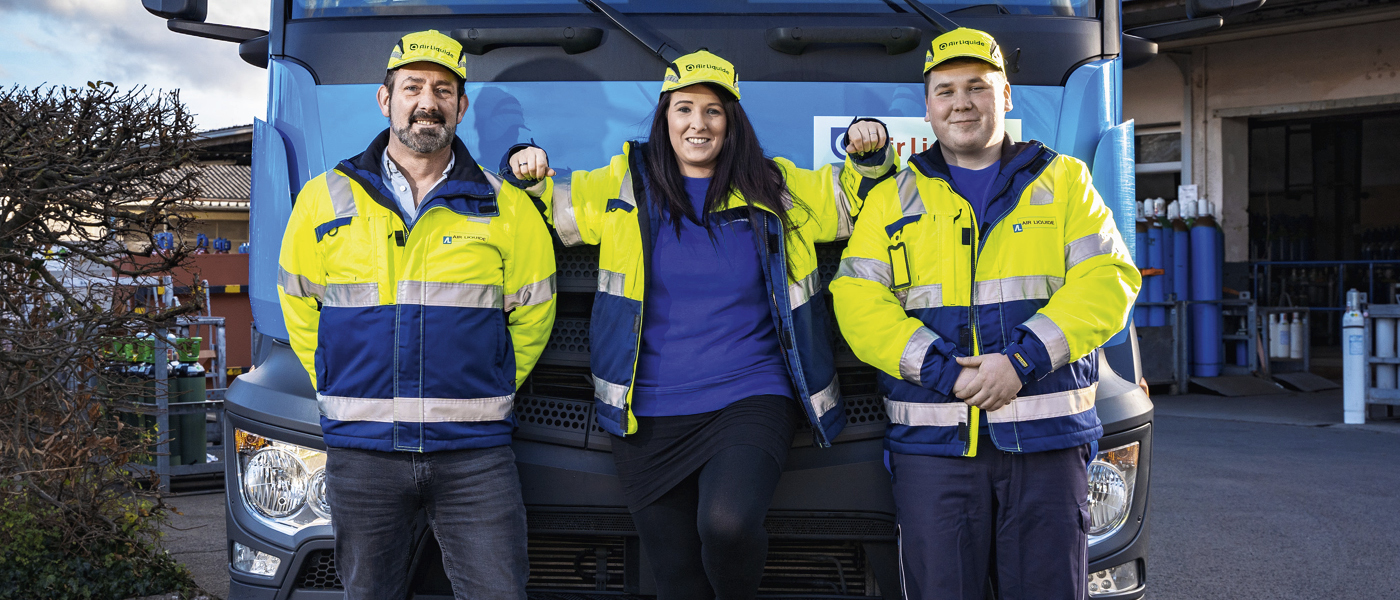 Drei Personen in gelb-blauen Warnjacken und gelben Mützen lehnen vor einem blauen Lkw mit deutschem Kennzeichen.