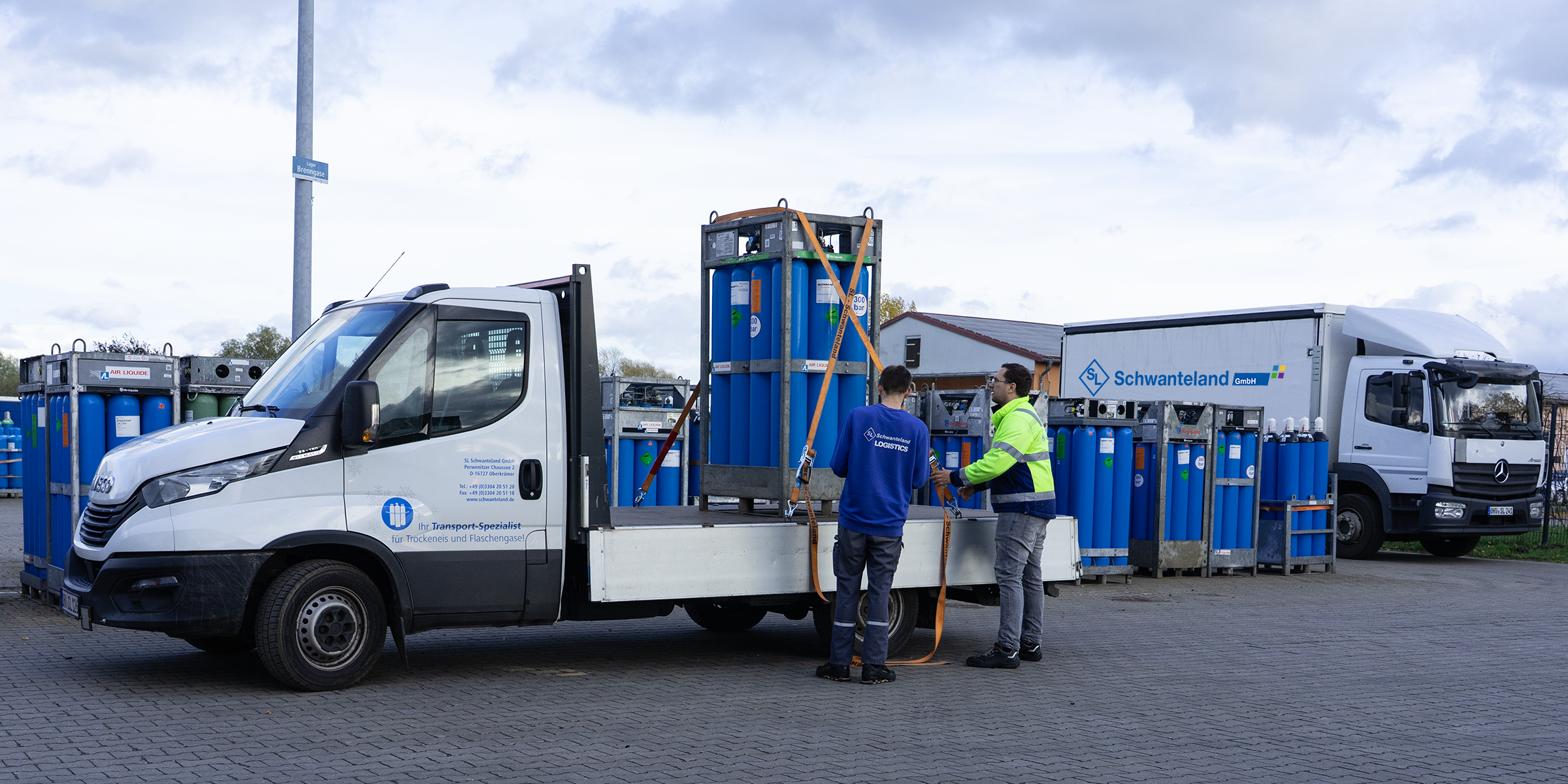 technische_gase_sl_schwanteland_header_v3 Zwei Männer sichern eine große blaue Gasflasche auf der Ladefläche eines weißen Transporters mit dem Schriftzug 'Schwanteland'. Im Hintergrund weitere blaue Gasflaschen und ein LKW mit 'Schwanteland'-Logo.
