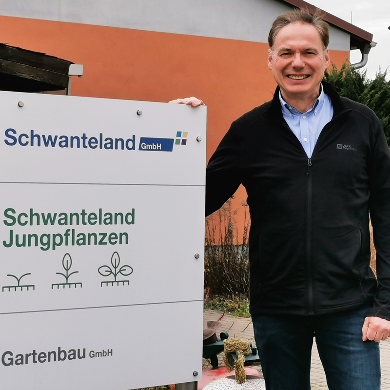 Mann in schwarzer Jacke steht neben Firmenschild mit den Namen 'Schwanteland GmbH', 'Schwanteland Jungpflanzen' und 'Gartenbau GmbH' sowie Pflanzensymbolen.