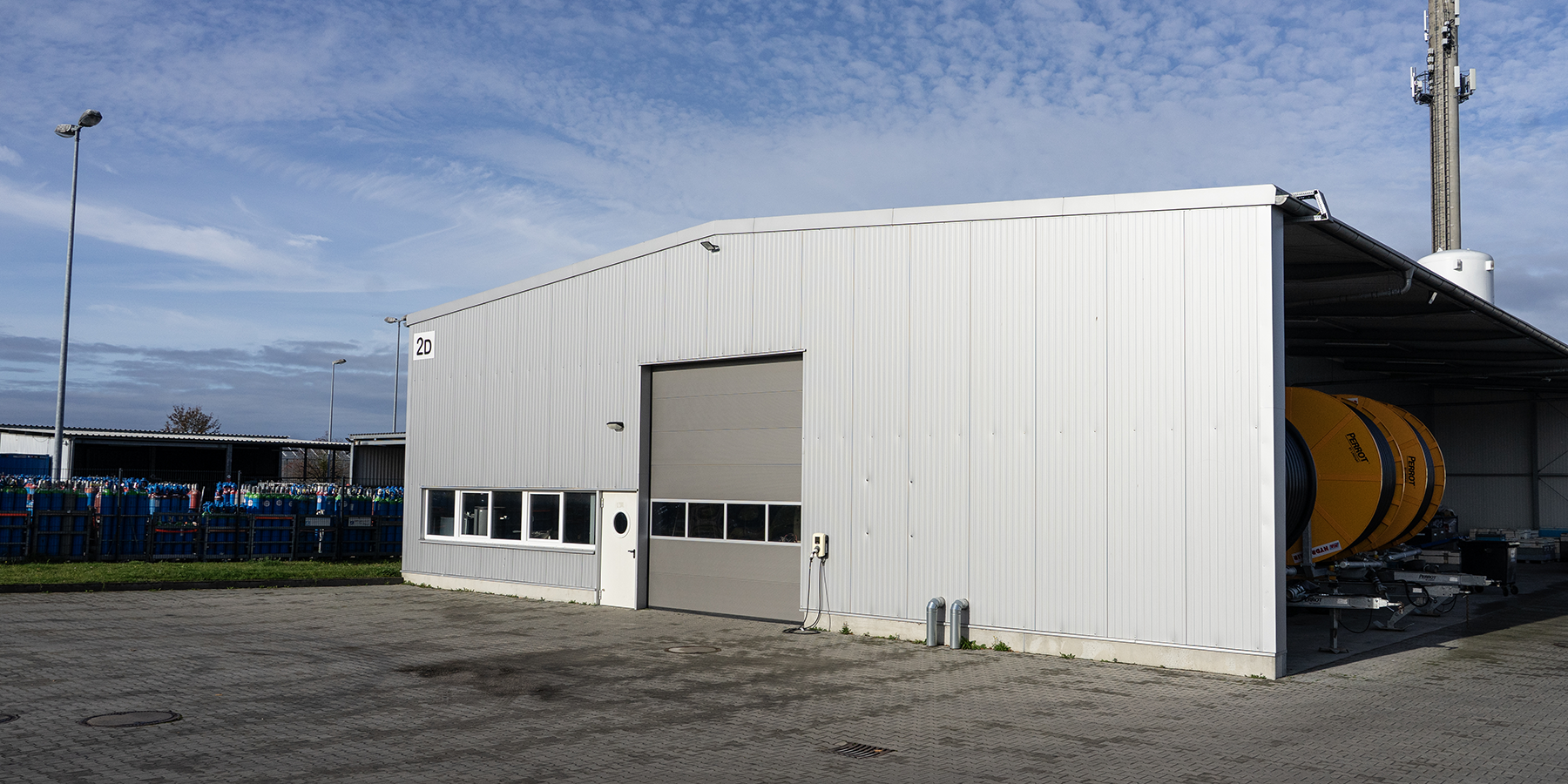 header_vermietung_sl_schwanteland_2 Große weiße Lagerhalle mit mehreren Fenstern und einem großen Rolltor, davor gepflasterter Hof unter blauem Himmel.