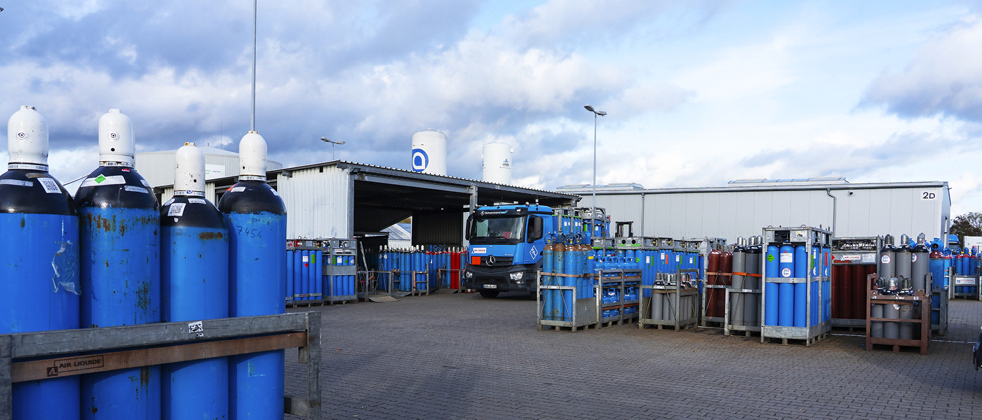 Außenbereich eines Gaseplatzes mit mehreren blauen Gasflaschen unterschiedlicher Größe, einem blauen Lkw und einem Gebäude unter bewölktem Himmel.
