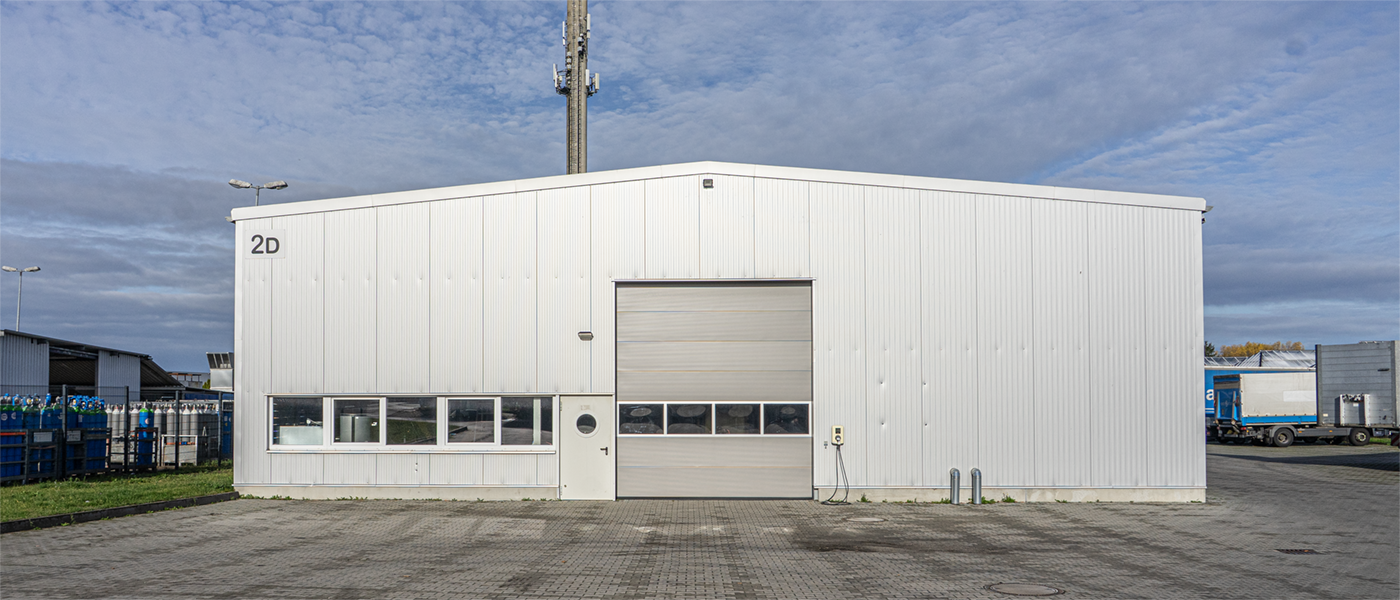 Große weiße Lagerhalle mit mehreren Fenstern und einem großen Rolltor, davor gepflasterter Hof unter blauem Himmel.