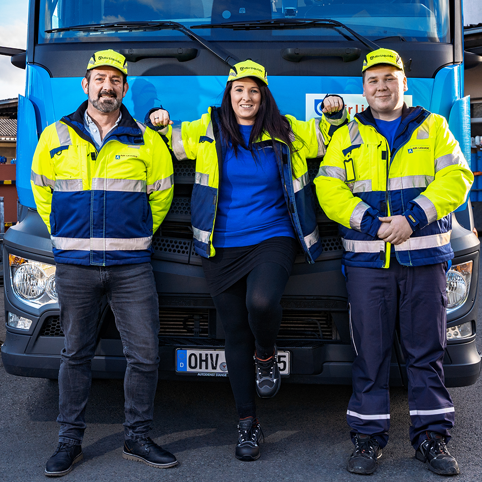 Drei Personen in gelb-blauen Warnjacken und gelben Mützen lehnen vor einem blauen Lkw mit deutschem Kennzeichen.