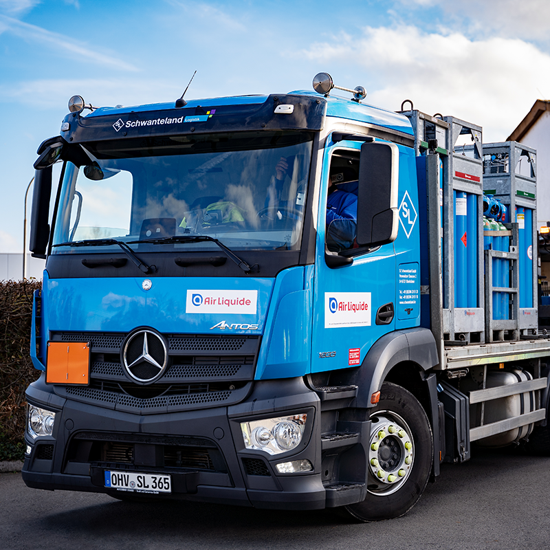 Blauer Mercedes-Lastwagen mit Aufschrift 'Schwanteland Logistik' transportiert mehrere Gasflaschen in Metallgestellen.