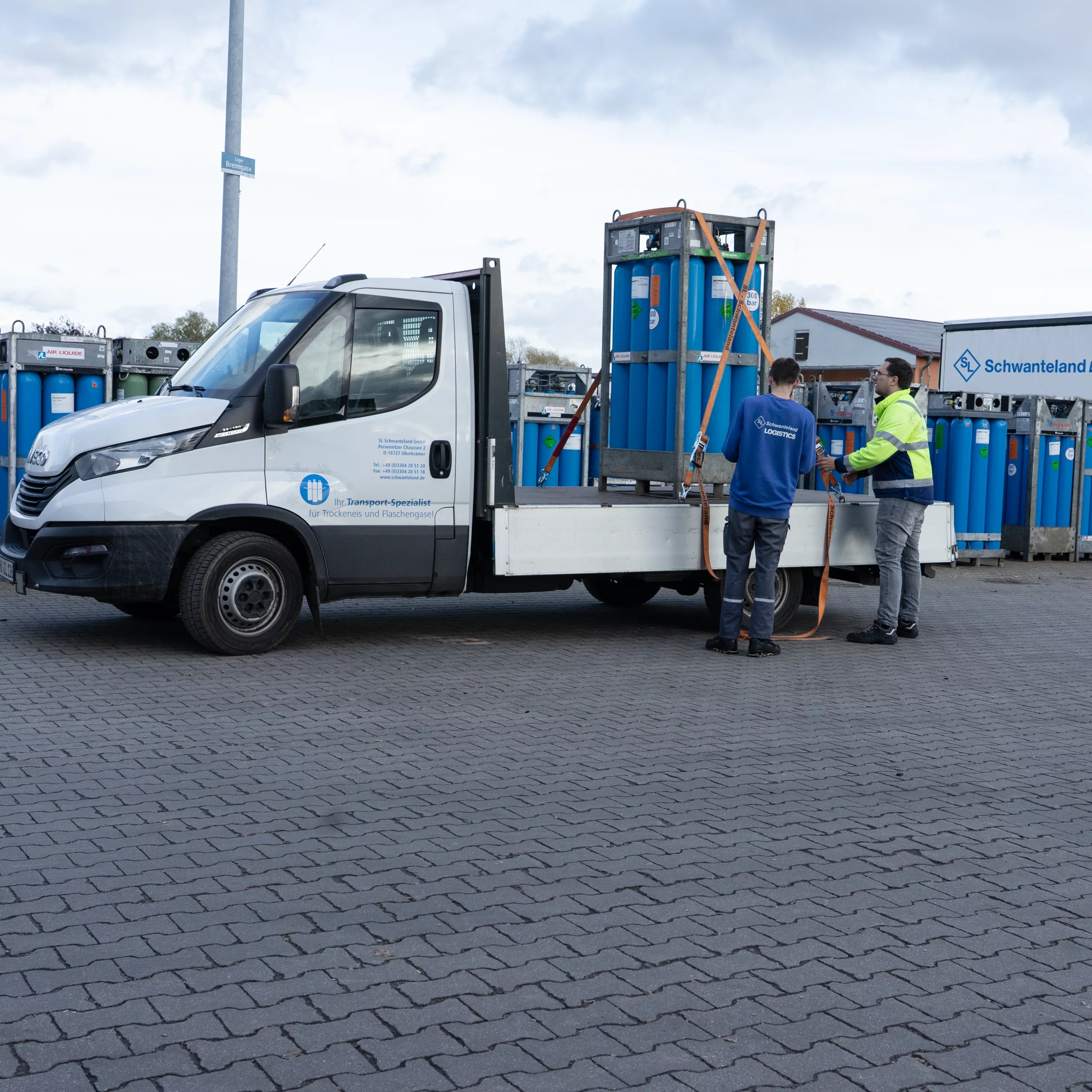 Zwei Männer sichern eine große blaue Gasflasche auf der Ladefläche eines weißen Transporters mit dem Schriftzug 'Schwanteland'. Im Hintergrund weitere blaue Gasflaschen und ein LKW mit 'Schwanteland'-Logo.