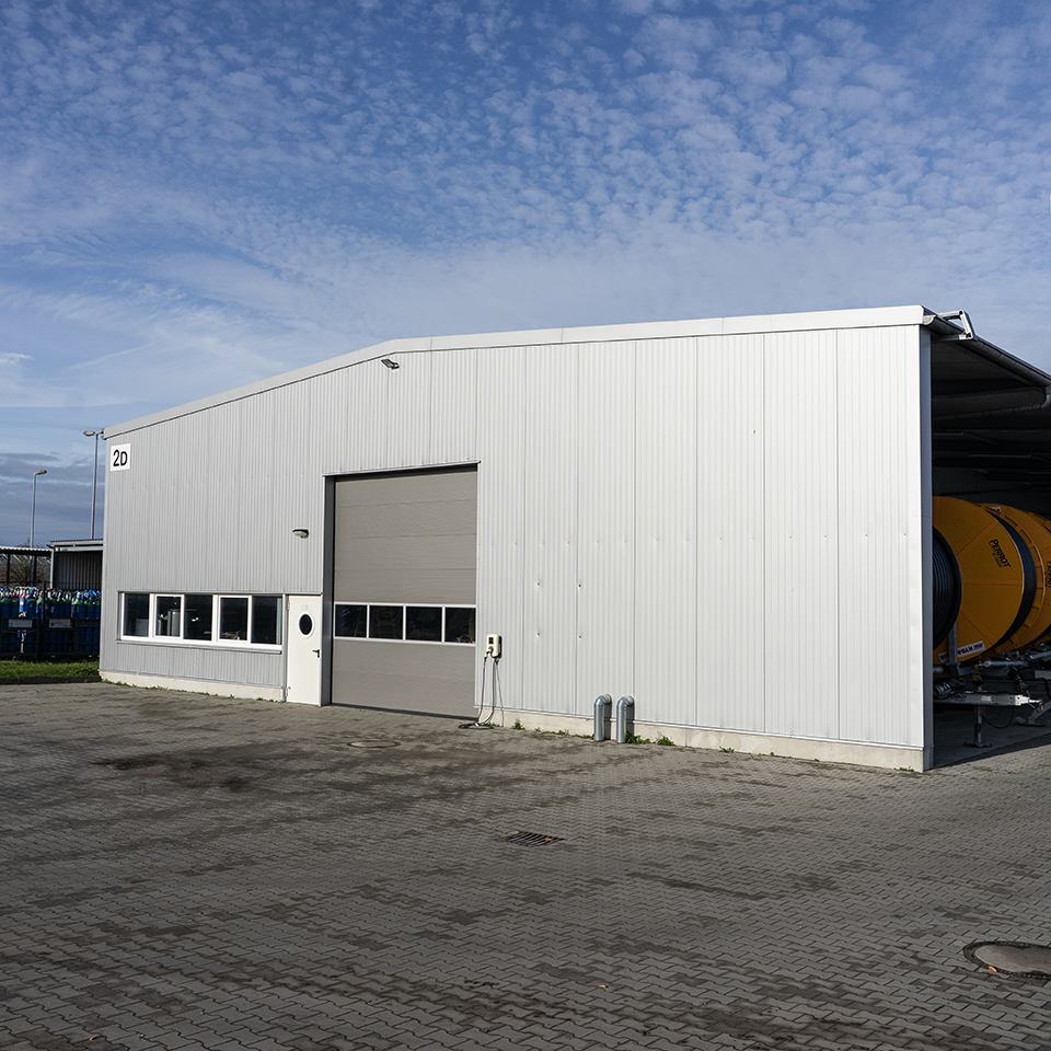 Große weiße Lagerhalle mit mehreren Fenstern und einem großen Rolltor, davor gepflasterter Hof unter blauem Himmel.