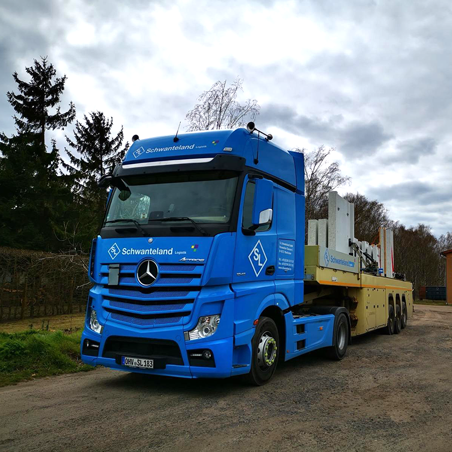 Blauer Mercedes-Lkw mit Aufschrift 'Schwanteland' transportiert mehrere große Betonfertigteile auf gelbem Anhänger auf unbefestigtem Weg.