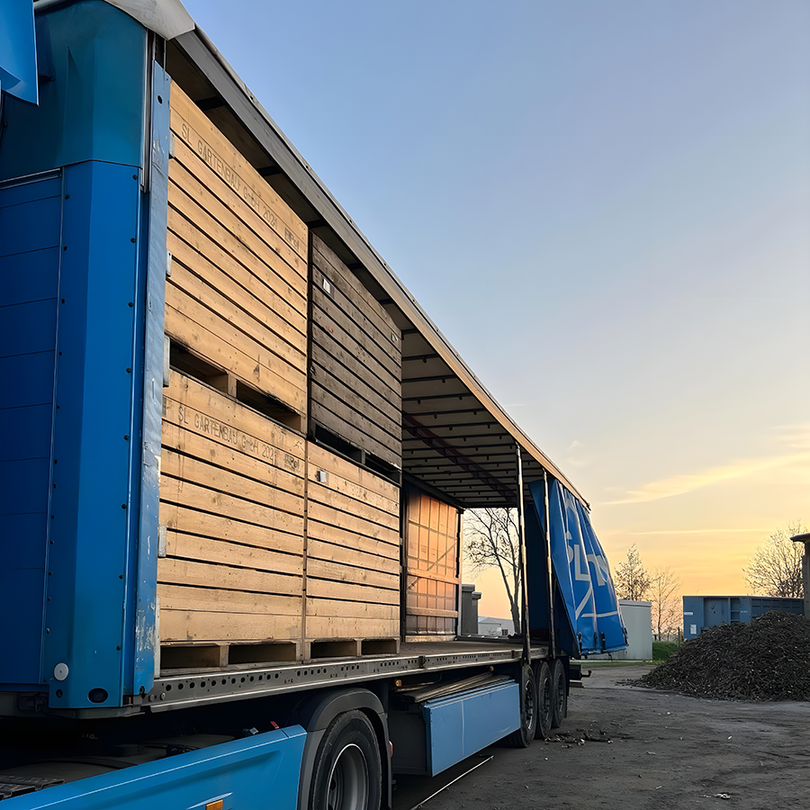 Blauer Lkw-Anhänger mit offener Plane, beladen mit gestapelten Holzkisten, im Sonnenuntergang auf einem Industriehof.