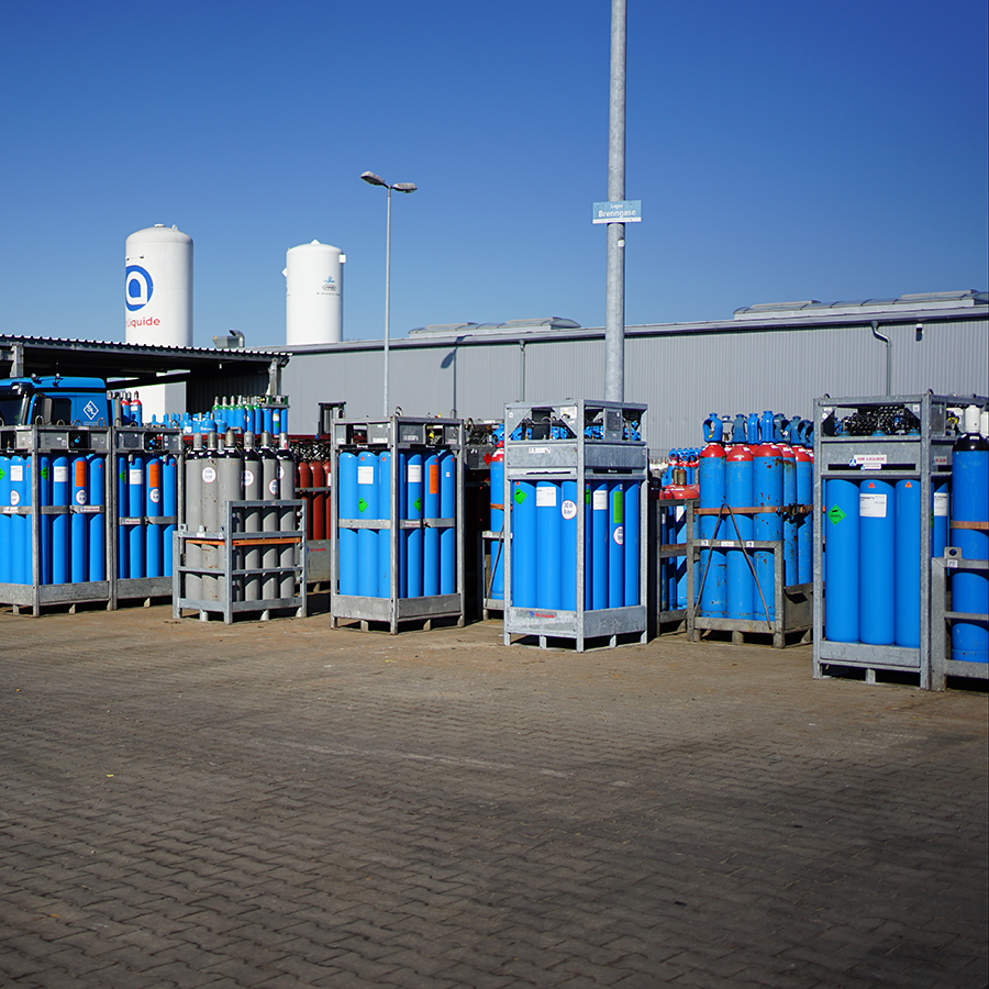 Mehrere Metallgestelle mit blauen Gasflaschen stehen auf einem gepflasterten Hof vor einer grauen Lagerhalle unter blauem Himmel.