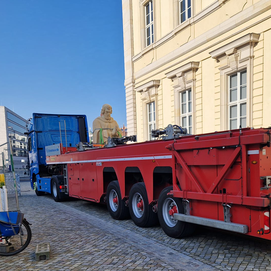 Blauer Sattelzug mit rotem Tieflader, darauf eine große Skulptur einer sitzenden Figur vor einem hellen Gebäude.