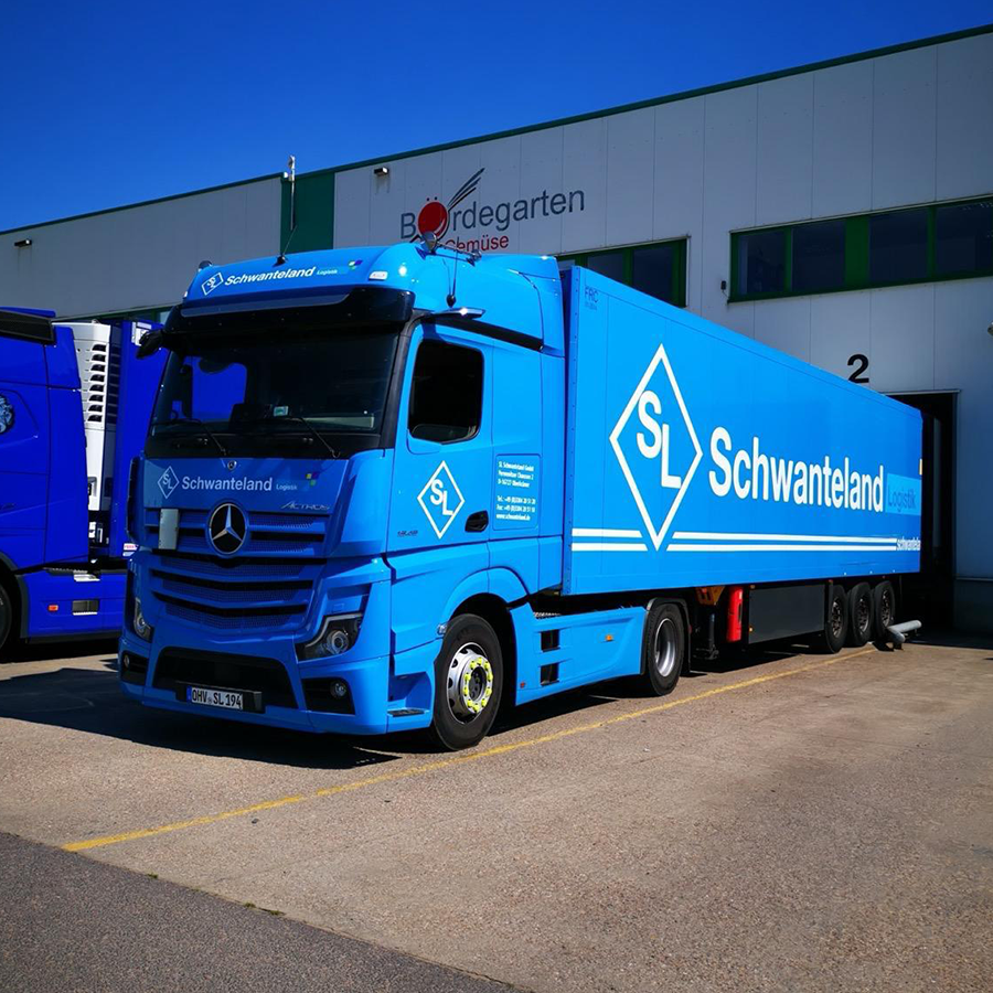 Blauer Sattelzug mit dem Schriftzug 'Schwanteland' und Logo, geparkt vor einem Gebäude mit dem Schriftzug 'Bordegarten Gemüse'.