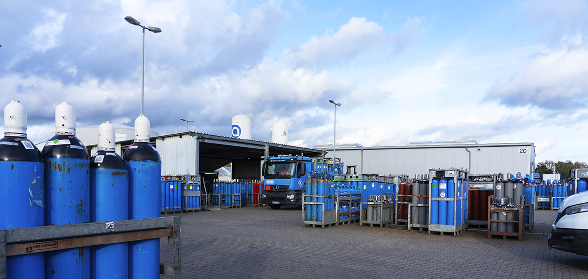 Außenbereich eines Lagers mit mehreren großen blauen und einigen roten Gasflaschen sowie einem blauen Lkw vor Lagerhallen unter bewölktem Himmel.