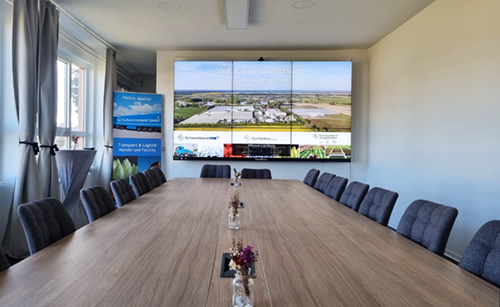 Moderner Seminarraum mit langem Holztisch, mehreren gepolsterten Stühlen und großer Leinwand mit Landschaftsaufnahme.