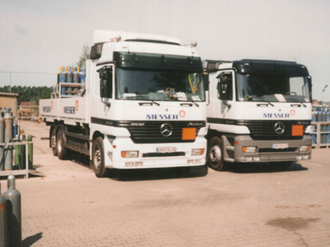 35_jahre_Schwanteland_beitrag_web2 Zwei weiße Mercedes-Lkw mit der Aufschrift "MESSER" stehen nebeneinander auf einem Hof, beladen mit Gasflaschen und Ausrüstung, historisches Bild aus den 90er Jahren.