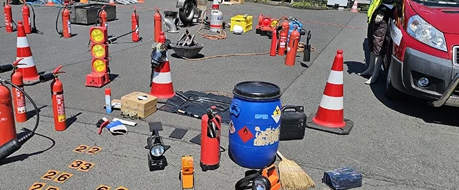 Mehrere Feuerlöscher, Warnkegel, Übungsmaterial und ein Feuerwehrfahrzeug stehen auf einem asphaltierten Platz, eine Person in Schutzkleidung und Helm steht rechts neben dem Fahrzeug.