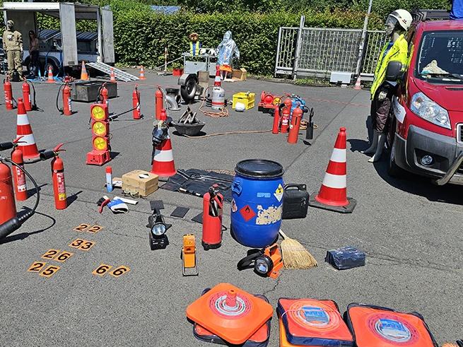Mehrere Feuerlöscher, Warnkegel, Übungsmaterial und ein Feuerwehrfahrzeug stehen auf einem asphaltierten Platz, eine Person in Schutzkleidung und Helm steht rechts neben dem Fahrzeug.